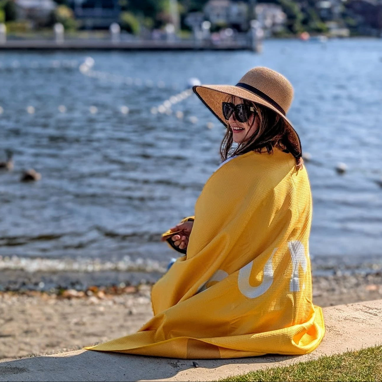 Person sitting by a ocean wearing a SUN'Y beach towel and wide-brimmed hat